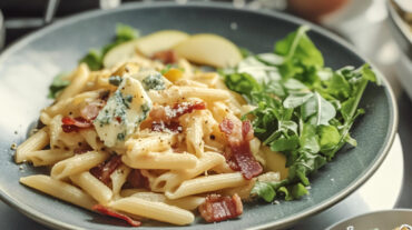 A photo of a pasta recipe on a plate containing penne, blue cheese and bacon bits